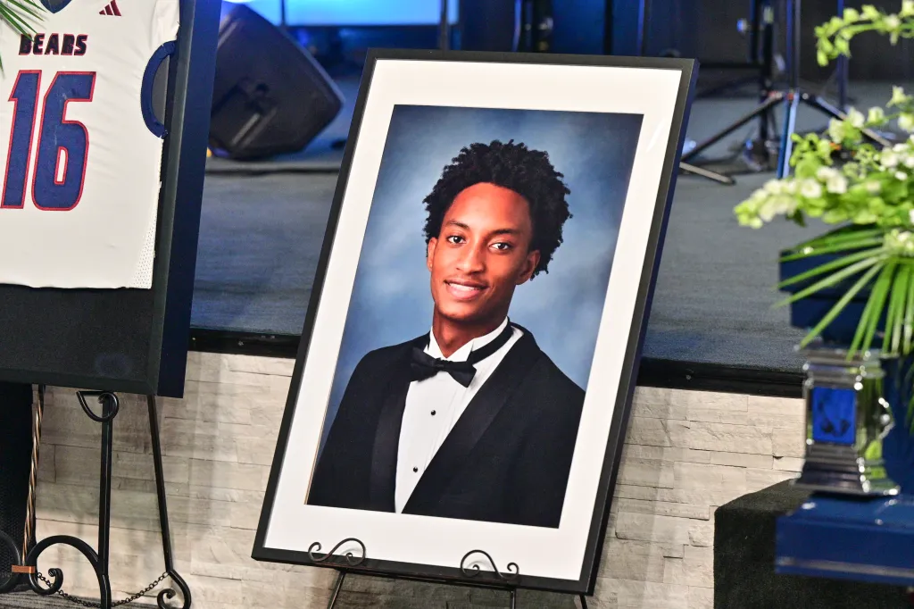 Funeral display with a pH๏τo of Nolan Wells in a tuxedo next to a white sports jersey with